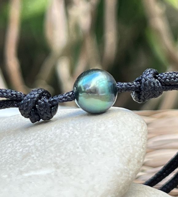 Une perle de culture de Tahiti ronde de 9,09 mm, couleur lagon aux reflets bleus et verts, de qualité AA et au lustre excellent, se révèle sur un lien macramé noir réglable
